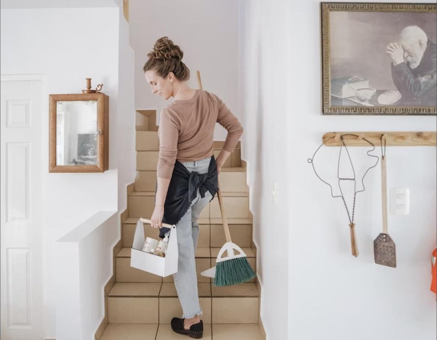 Cleaner carrying a caddy of supplies and a broom through a bright, tidy home